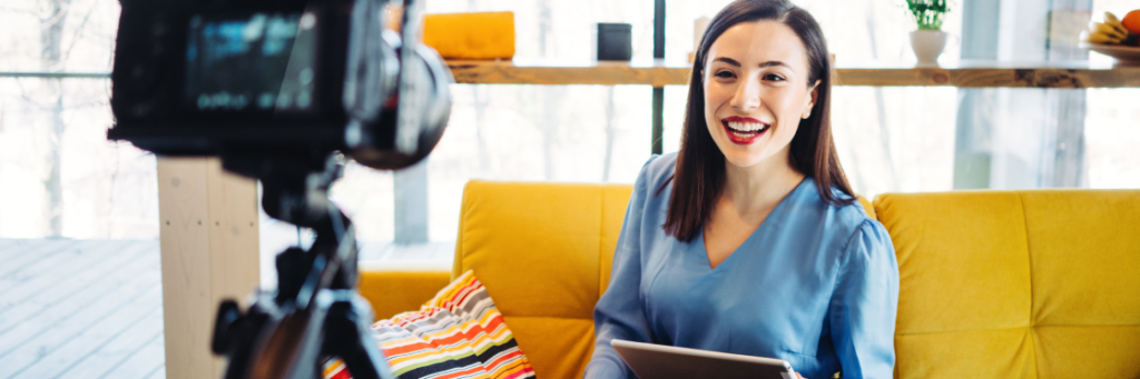 A smiling woman in a blue top sitting on a yellow sofa holding a tablet, facing a Biteable recording camera in a bright, modern room.