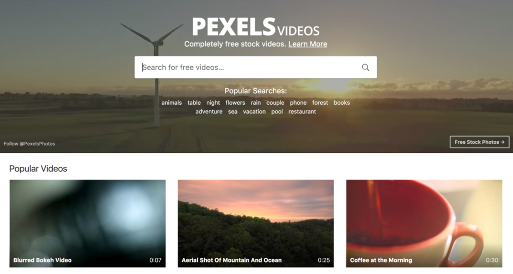 Website homepage for pexels videos featuring a search bar and thumbnails for popular videos, including a blurred bokeh video, an aerial shot of a mountain and ocean, and a coffee cup.