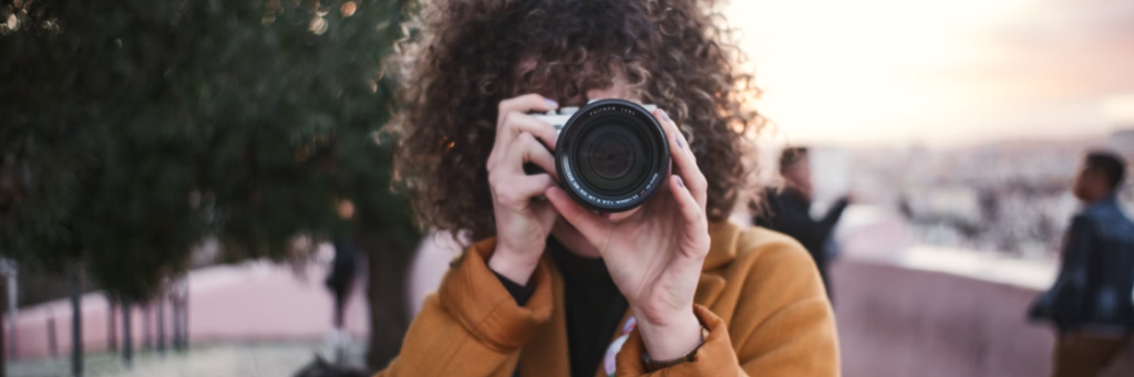 Person with curly hair holding a camera up to their face, taking a photo at sunset.