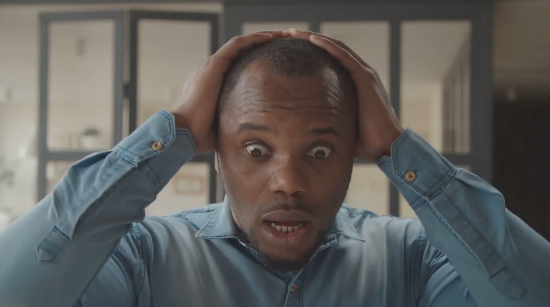 A man in a blue shirt looking shocked with his hands on his head in a modern room, creating content with Biteable video maker.