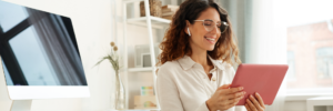 Smiling woman wearing glasses using a tablet in a modern office with a desktop computer in the background.