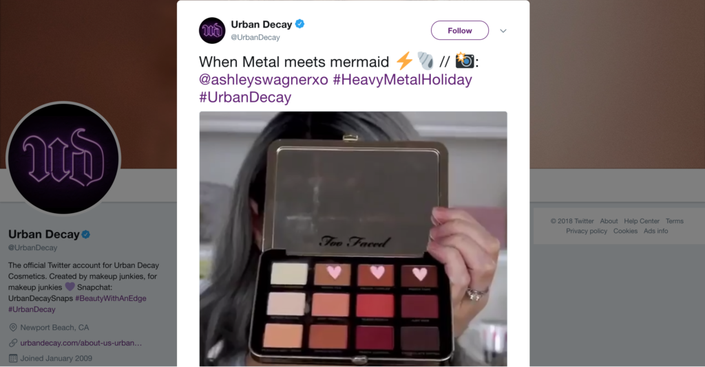 A person holding an open too faced makeup palette, with a tweet from urban decay promoting a metallic eyeshadow look with hashtags referencing a collaboration and a holiday theme.