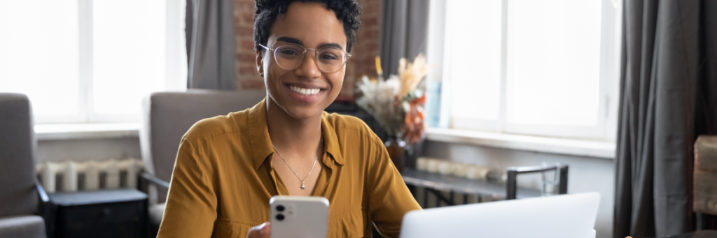 Woman in a yellow blouse smiling while using a smartphone with a laptop in front of her.