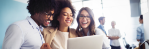 Three professionals smiling and looking at a laptop screen in a modern office setting.