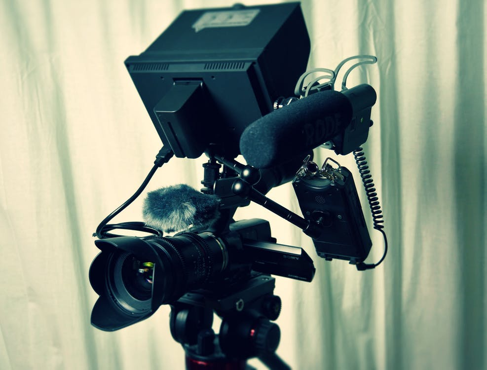 Professional video camera with attached microphone and accessories on a tripod against a draped white background.