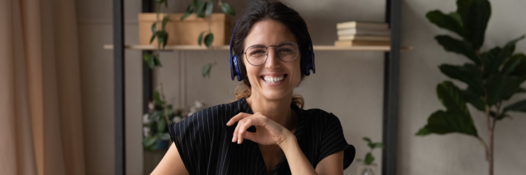 A smiling woman with glasses and headphones sits at a desk in a modern office, using Biteable video maker, with books and plants in the background.