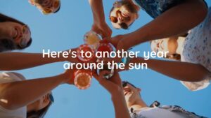 A group of friends toasting with cocktails under a clear blue sky, viewed from below, with celebratory text "here's to another year around the sun," created using Biteable video maker.