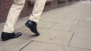 A person walking on a paved sidewalk, with focus on their black shoes and the lower part of their light-colored pants captured in a Biteable video.