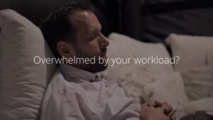 Man lying on a bed, appearing stressed, with text overlay reading "Overwhelmed by your workload? Try Biteable video maker.