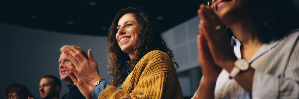 Diverse group of people clapping enthusiastically in an auditorium, focusing on a smiling woman in a yellow sweater featured in a Biteable video.
