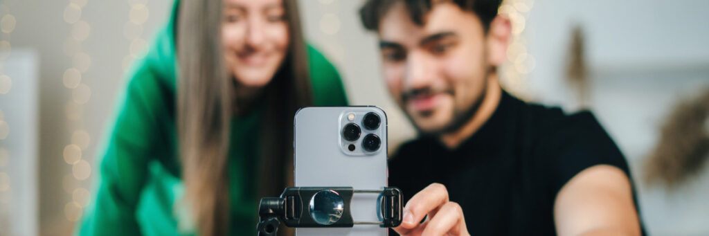 A young couple capturing a selfie with a smartphone mounted on a tripod, Biteable video maker and blurred Christmas lights in the background.
