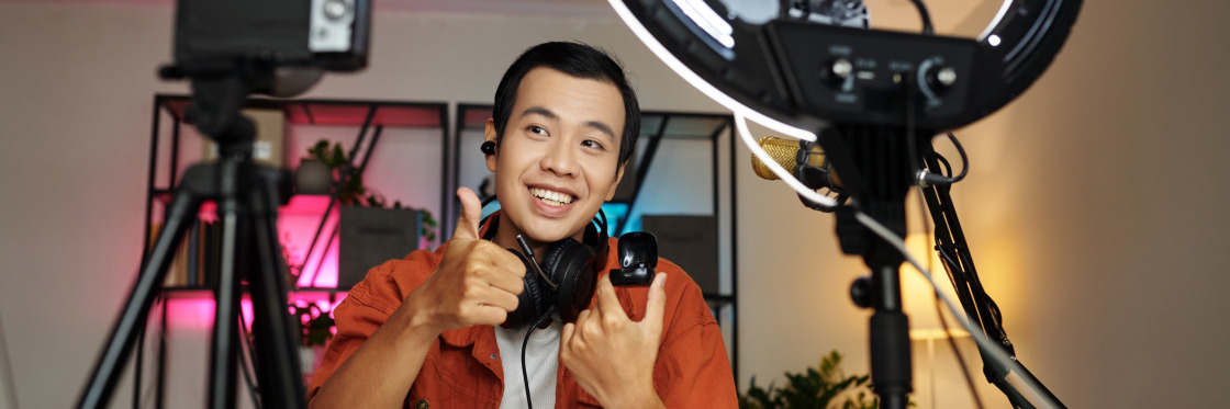 Young man holding a Biteable video maker camera, giving a thumbs up between two ring lights and tripods, with a pleasant expression in a well-lit room.