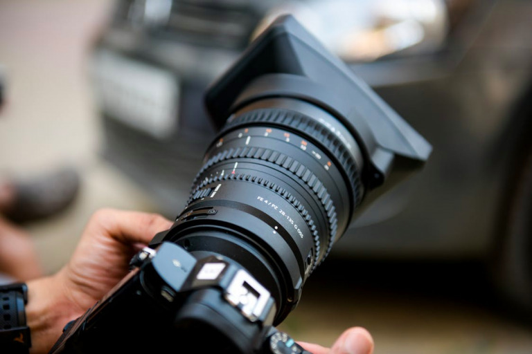 Close-up of hands adjusting a large camera lens with a car blurred in the background, captured using Biteable video maker.