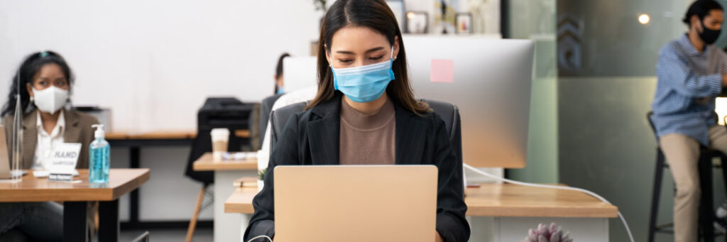 Woman wearing a mask works on a laptop using Biteable video maker in a socially distanced office setting with colleagues in the background.