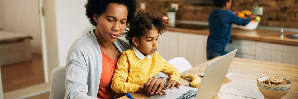 A mother and her young daughter sit at a kitchen table, using a Biteable video maker on a laptop together, while a boy plays in the background.