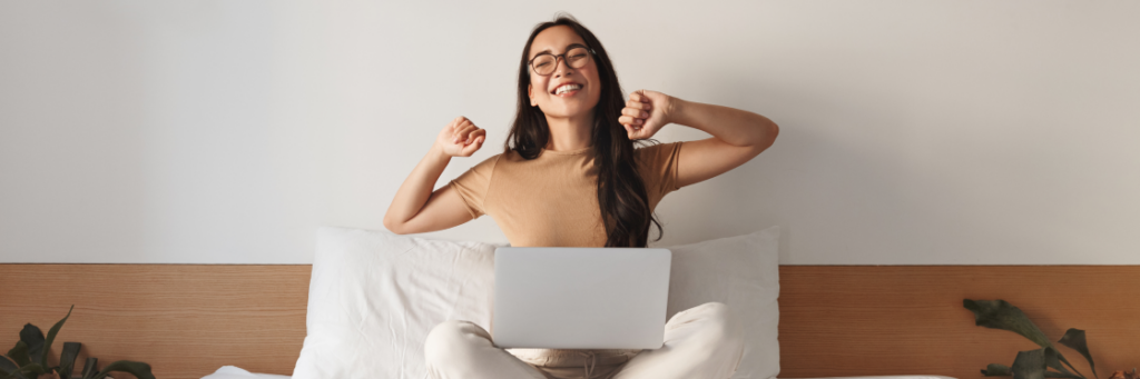 Young woman in glasses sitting on a bed with a laptop, joyfully stretching her arms with a pleased expression, embodies the flexible work future.