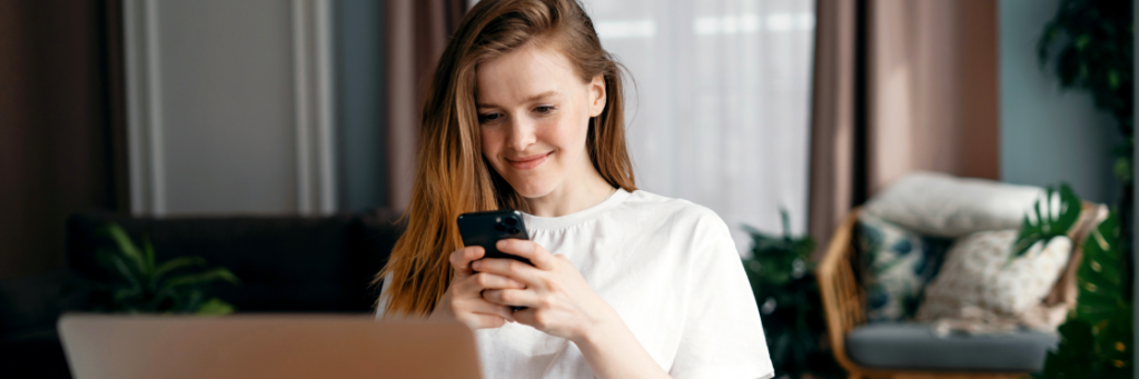 Young woman using a smartphone for social media at a desk with a laptop and indoor plants in the background.