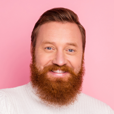 Man with a red beard smiling, wearing a white sweater, against a pink background.