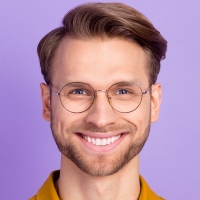 Close-up of a smiling man with neat hair wearing round glasses and a mustard-colored shirt against a purple background.