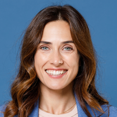 Portrait of a smiling Caucasian woman with long brown hair, wearing a blue blazer, against a blue background.