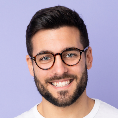 A portrait of a smiling man with dark hair and beard, wearing glasses and a white T-shirt against a purple background.