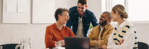 Four colleagues laugh and discuss around a laptop in a bright office setting, conveying a collaborative and cheerful work environment.