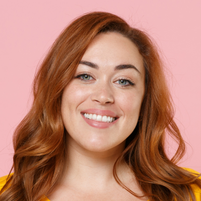 A smiling woman with long reddish-brown hair wearing a yellow top against a pink background.