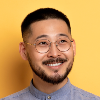 Portrait of a smiling Asian man with a beard and glasses, wearing a blue shirt against a yellow background.