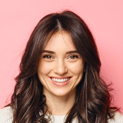 A woman with long, wavy brown hair smiling at the camera, against a pink background.