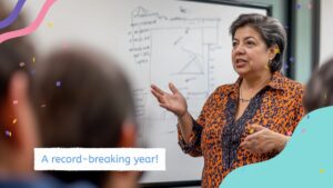 A woman in an orange patterned blouse speaks to an audience in front of a whiteboard, with text overlay reading "A record-breaking year!.