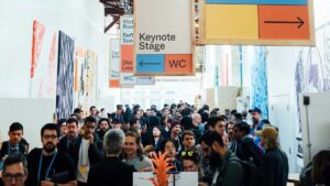 A large group of people gathers in a hallway at a conference, with directional signs for the keynote stage and restrooms hanging overhead.