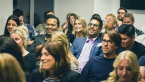 A group of people sit closely together in a brightly lit room, attentively listening and smiling during an event or presentation.