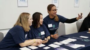 Three women wearing "Product School" shirts sit at a table with name tags and papers while one takes a selfie with her phone.