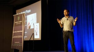 A man stands on stage presenting at a conference, with a projected slide and a banner reading "Product School Silicon Valley" visible beside him.