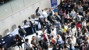 Large group of people gather at a busy indoor event with information tables and posters along the wall.