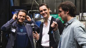 Three men stand indoors holding microphones, one wearing a headset and adjusting it. Various electronic equipment and cables are visible in the background.