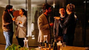 Six people stand in small groups at an indoor social gathering, conversing and holding drinks, with bottles and glasses on a table in the foreground.