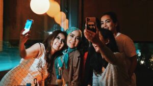 A group of five women pose closely together and smile while taking selfies with their smartphones in a dimly lit indoor setting.