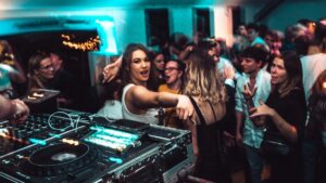 A crowded party scene with a DJ setup in the foreground and people dancing and socializing under colorful lights.