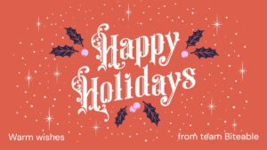 Red holiday card with white ornate text reading "Happy Holidays," decorated with holly leaves and berries. Smaller text at bottom says, "Warm wishes from team Biteable.