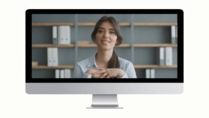 A woman sits in front of shelves and looks at the camera during a video call, displayed on a computer monitor.