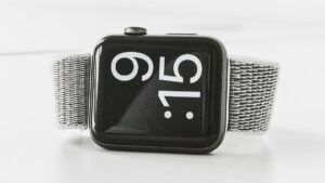 A close-up of a smartwatch with a gray fabric band displaying the time 9:15 in large white numerals on its screen.