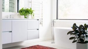 Modern bathroom with a white floating vanity, freestanding tub, large window, indoor plants, and a red patterned rug on the floor.