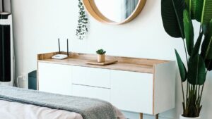 A wooden sideboard with a round mirror above it, a potted plant, and a Wi-Fi router on top, next to a large leafy houseplant in a bright, modern bedroom.