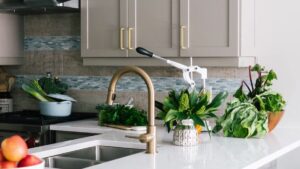 Modern kitchen with a gold faucet, gray cabinets, a produce juicer, and various fresh vegetables and greens arranged on the white countertop.