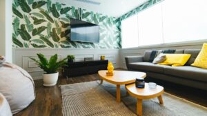 Modern living room with a leafy wallpaper accent wall, wall-mounted TV, gray sofa with yellow cushions, potted plant, and wooden coffee table on a striped rug.