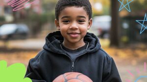 A young boy in a black hoodie holds a basketball and smiles outdoors, with colorful doodles around the image.