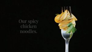 A fork holds a swirl of noodles garnished with parsley against a black background, with the text "Our spicy chicken noodles.