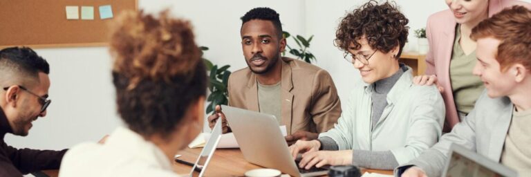 A group of people sit around a table in an office, having a discussion while using laptops and documents.