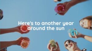 People holding colorful drinks in a circle, viewed from below against a blue sky, with the text "Here&rsquo;s to another year around the sun" in the center.